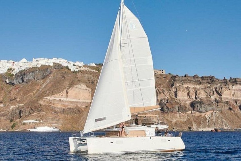 Un velero con vela blanca navegando en aguas tranquilas, una costa rocosa y edificios blancos al fondo. 3