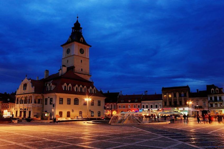 Ein historisches Gebäude mit einem Glockenturm, ein Platz mit einem Brunnen und umgebende Gebäude. 2