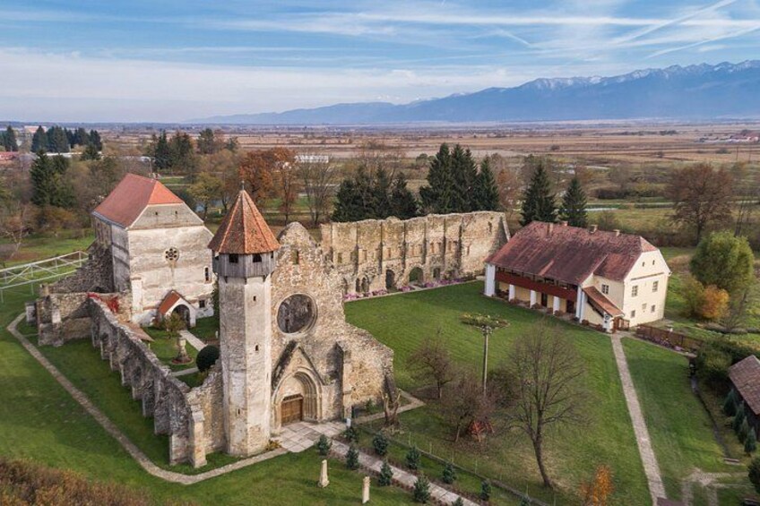An ancient stone church with a bell tower, a white house with a red roof, and a grassy field with trees and mountains in the background. 4