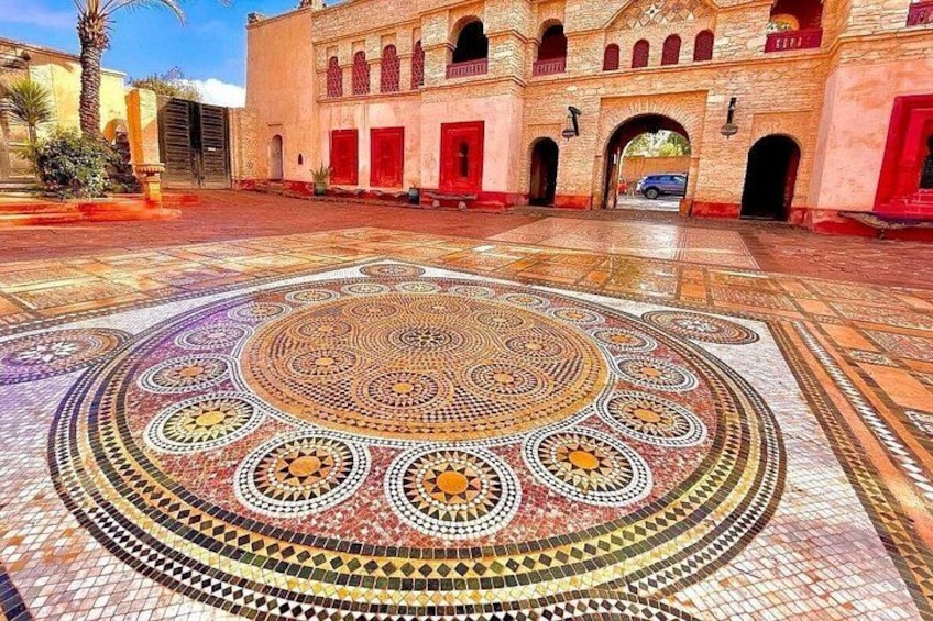 A courtyard with intricate mosaic flooring and a building with arched windows and red doors. 2