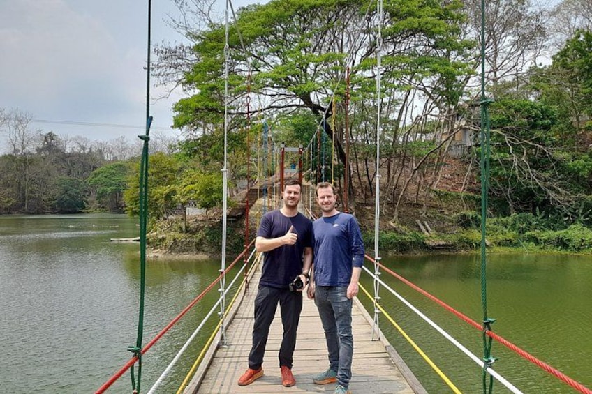 Two people standing on a suspension bridge over a river, surrounded by trees and greenery. 5