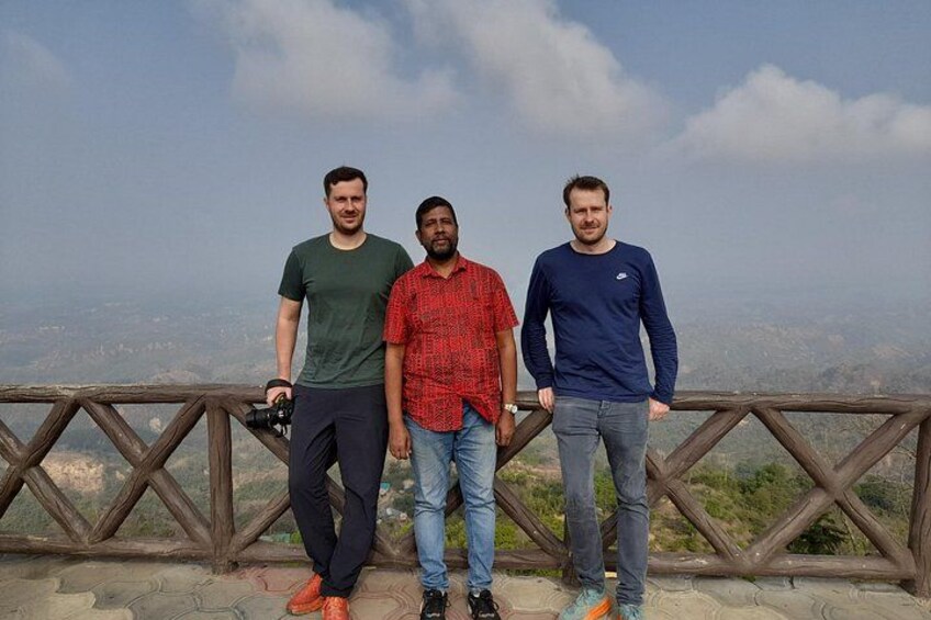 Three people standing on a wooden platform with a scenic view of a mountainous landscape. 2