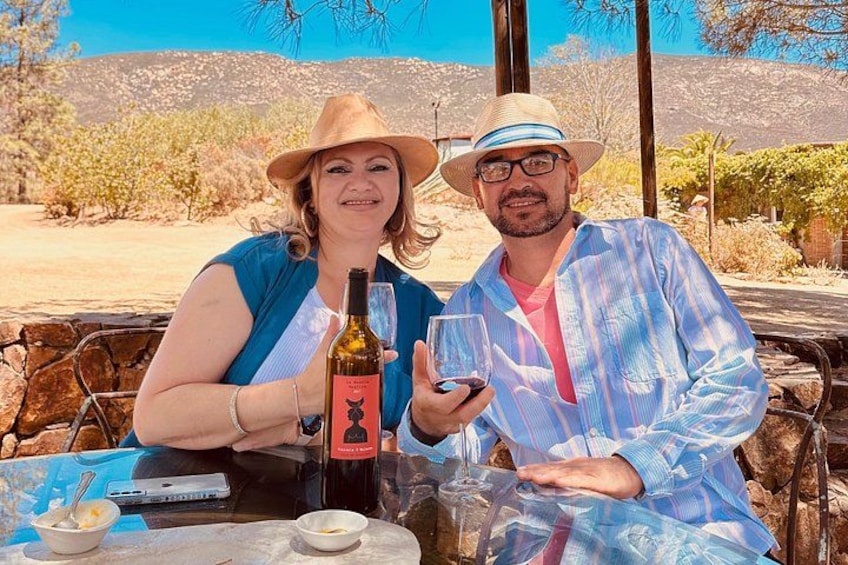 Two people sitting at a table outdoors with a bottle of wine and glasses. 2