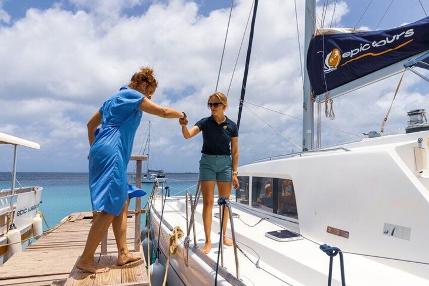 Twee mensen op het dek van een zeilboot, één in een blauwe jurk en de ander in een zwart shirt en korte broek, houden elkaars hand vast. 2