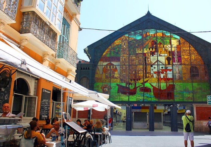 Un mural colorido en un edificio, mesas al aire libre en un café y una persona parada en la acera. 1