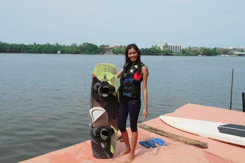 Een persoon staat op een steiger met een kajak en een paddleboard, een rustig wateroppervlak en een verre oever met gebouwen. 4