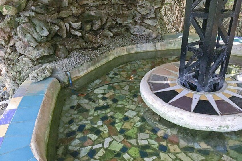 Ein mit Mosaikfliesen ausgelegter Brunnen mit einer Steinwand und einer Metallkonstruktion in der Mitte. 2