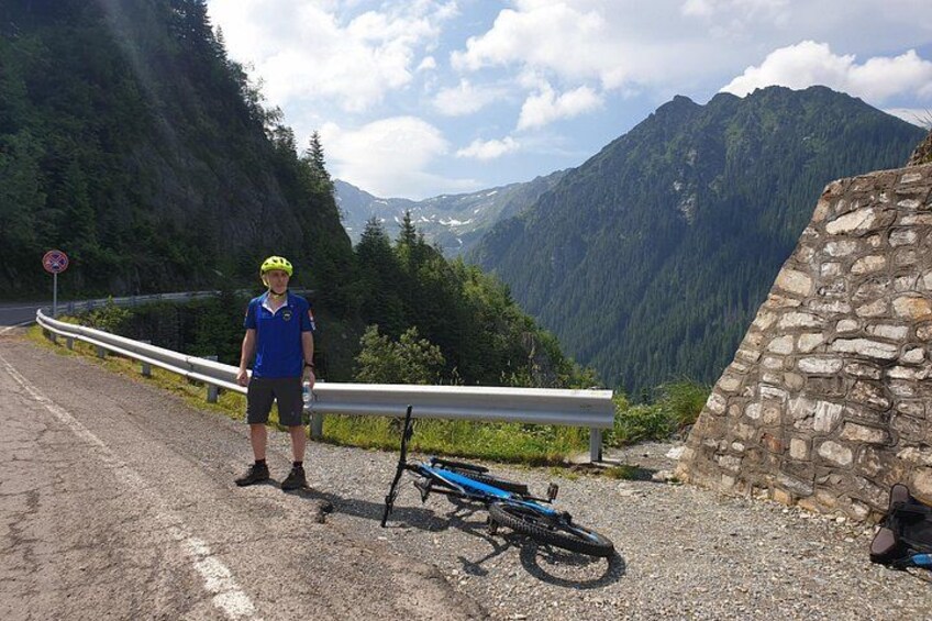 A cyclist standing next to a bicycle on a mountain road with a stone wall and forested mountains in the background. 3