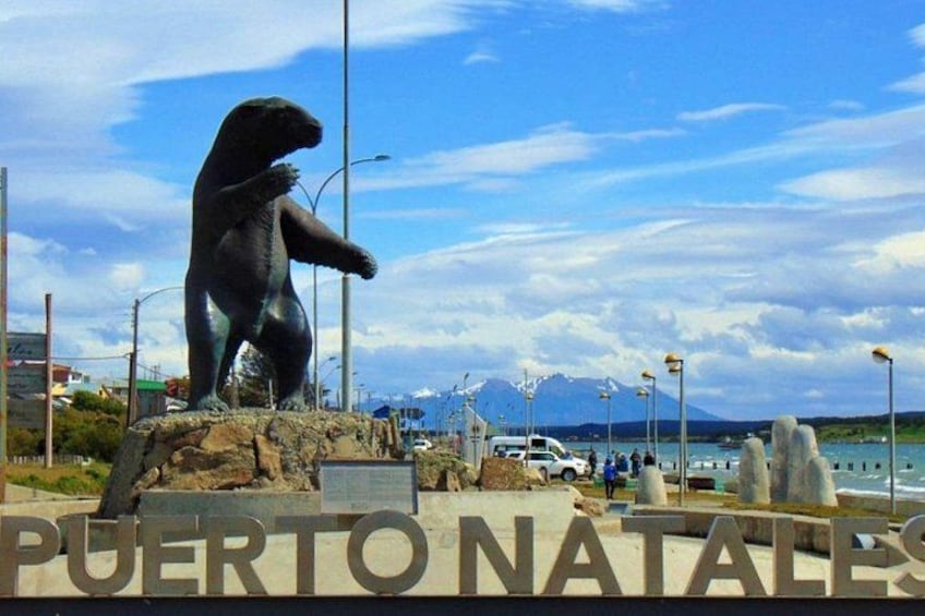 A bear statue, a sign reading 'PUERTO NATAL,' and a body of water with mountains in the background. 2