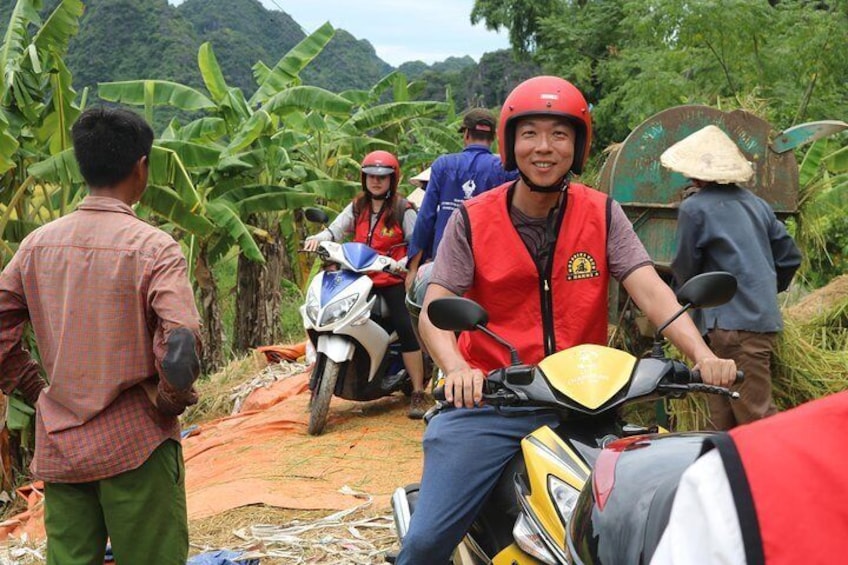 People on motorcycles in a rural area with banana trees and a person carrying a basket. 5