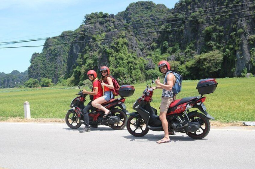 Three people on motorcycles on a road with a green landscape and hills in the background. 3