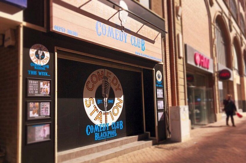 A Comedy Club storefront with a sign, a window display, and a person walking in the background. 1