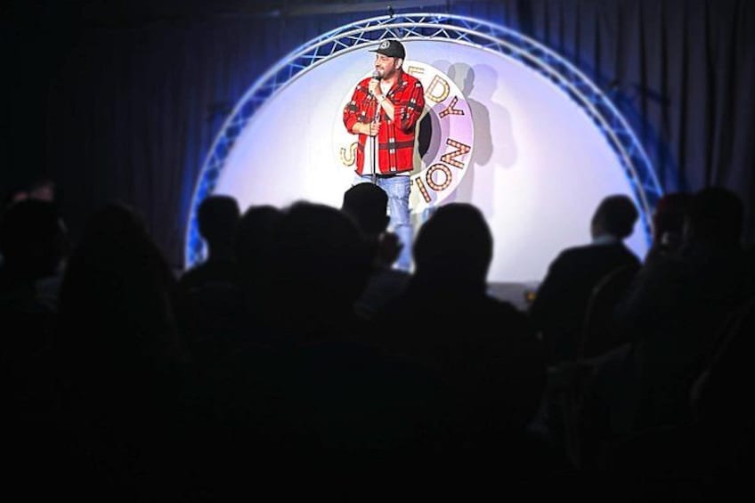 A person on stage with a microphone, audience in front, and a large illuminated sign with text. 2