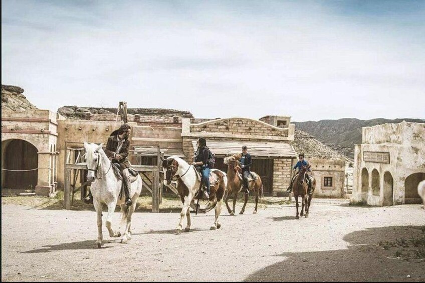 A group of people riding horses in a dusty, open area with old buildings in the background. 4