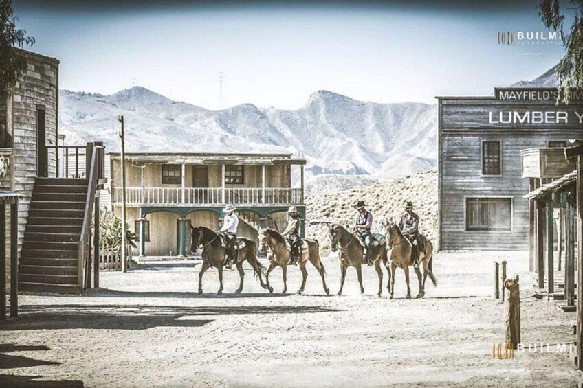 A group of people on horseback in a desert town with buildings and mountains in the background. 1