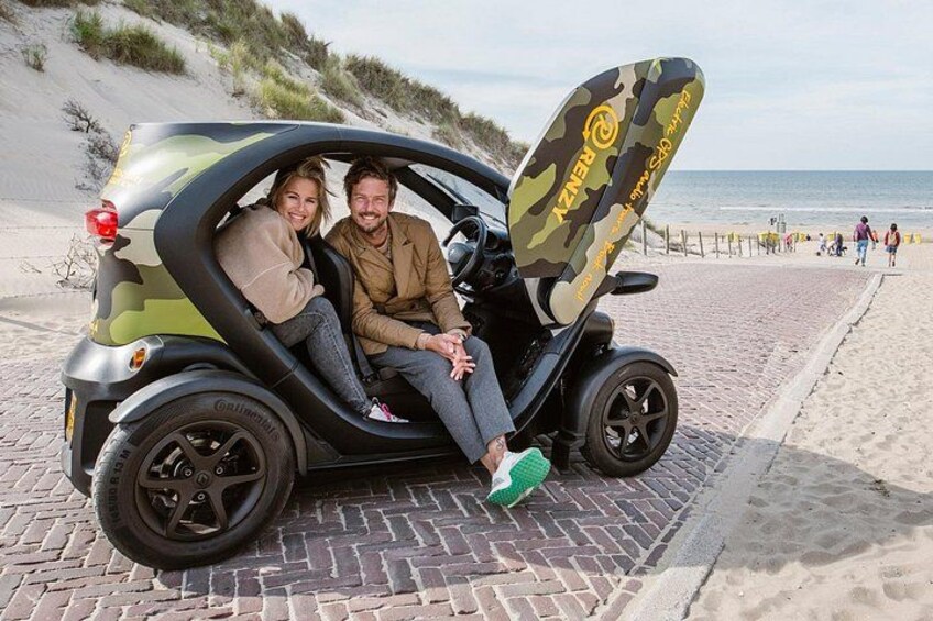 Una coppia in un piccolo veicolo scoperto con motivo mimetico sul fianco, parcheggiata su un vialetto asfaltato vicino a una spiaggia. 1