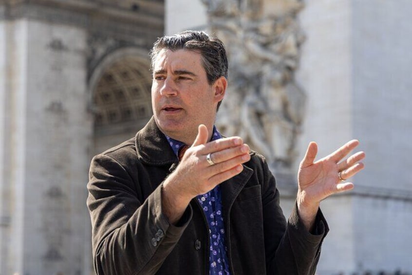 A man in a coat gesturing in front of a historic building with detailed architecture. 1
