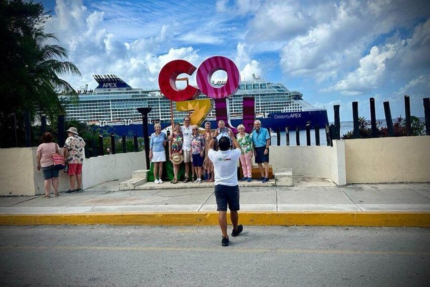 A group of people standing in front of a large sign with the letters 'GO' and a cruise ship in the background. 3