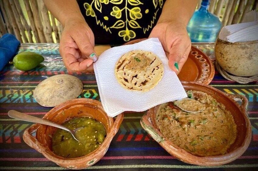 En person holder en tortilla med en ske grøn sauce og en skål brun sauce på en farverig dug. 5