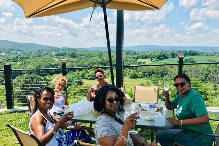 A group of people sitting at an outdoor table with a scenic view of a green landscape and hills in the background. 2