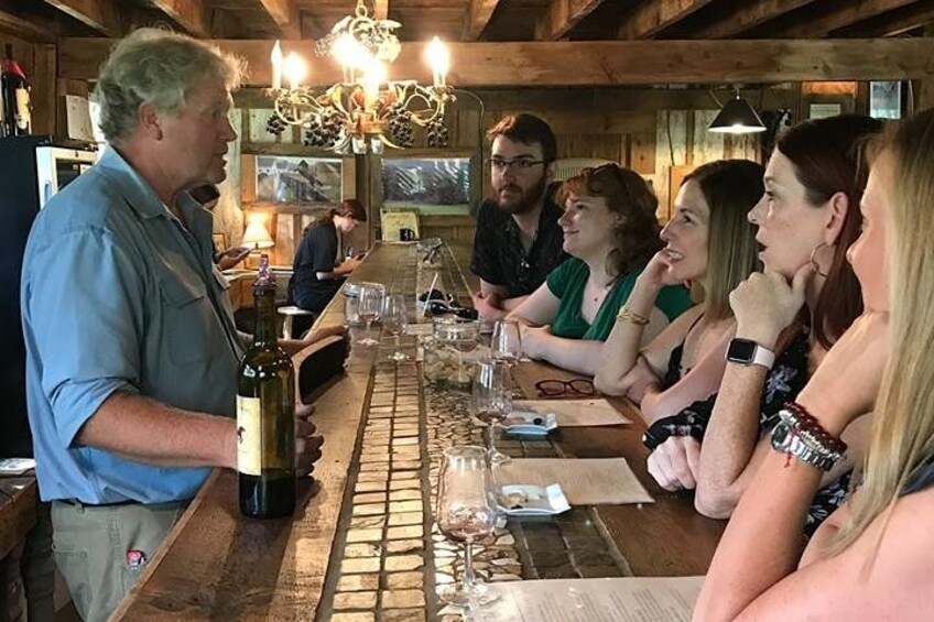 A group of people seated at a long wooden table in a rustic setting with a bar counter, wine glasses, and a bottle of wine. 2