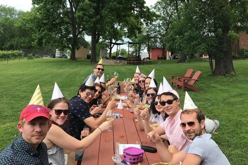 A group of people toasting at a wooden table outdoors with trees and a grassy lawn in the background. 4