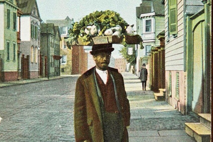 A man carrying a large load of vegetables on his head, walking down a street lined with buildings. 2