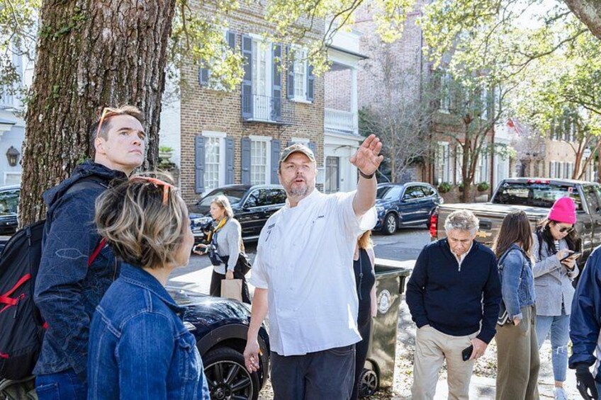 A group of people standing on a sidewalk, a tree, and parked cars. 4