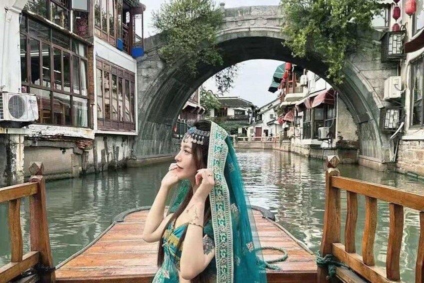A person on a wooden boat in a canal with traditional buildings and a stone bridge in the background. 1