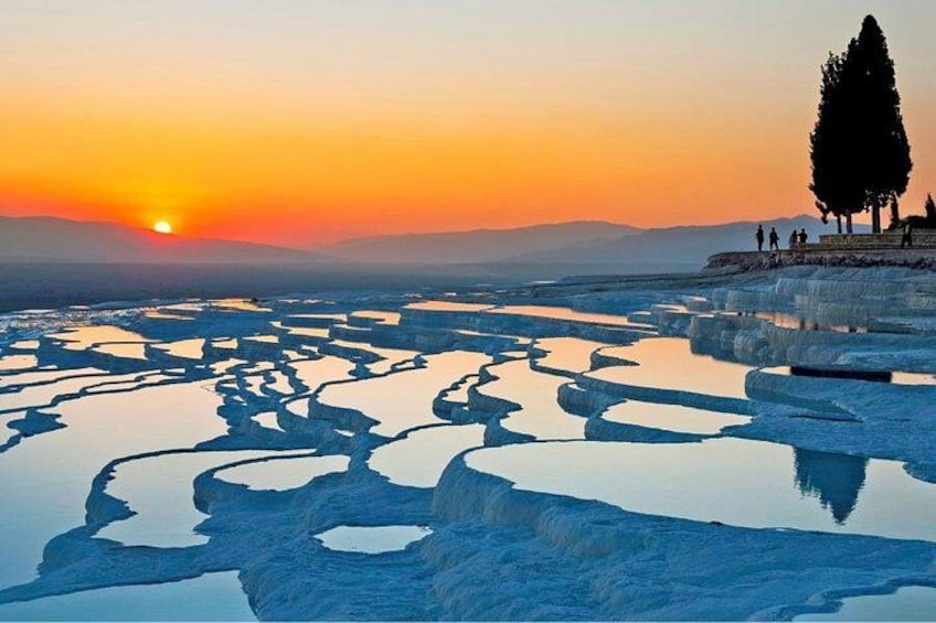 A sunset over a salt flat with reflective pools, a lone tree, and a group of people on a viewing platform. 2