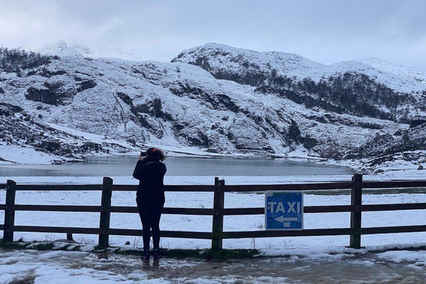 A person taking a photo near a lake with snow-covered mountains in the background and a sign that reads 'TAXI' on a wooden fence. 2