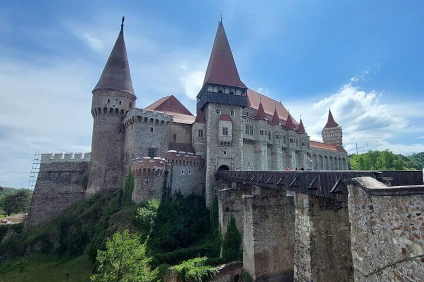 A historic castle with multiple towers and a red roof, surrounded by greenery and a clear sky. 1
