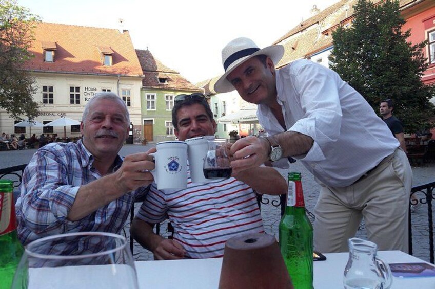 Three people toasting with mugs at an outdoor table with bottles and glasses. 3
