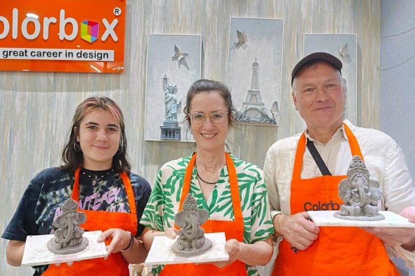Tres personas sostienen esculturas con delantales anaranjados frente a una pared con arte y un letrero que dice 'colorbox gran carrera en diseño'. 4