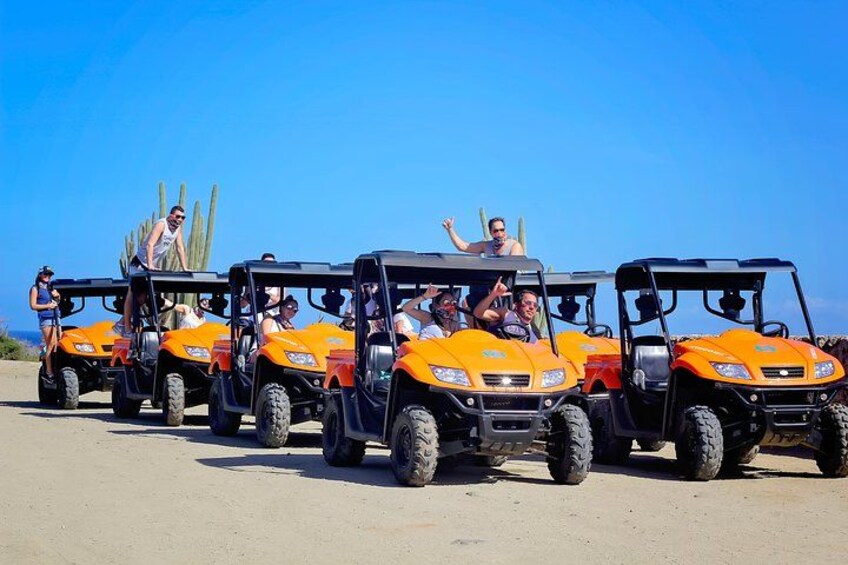 Eine Reihe orangener Geländewagen mit Passagieren, abgestellt auf einer unbefestigten Straße. 1