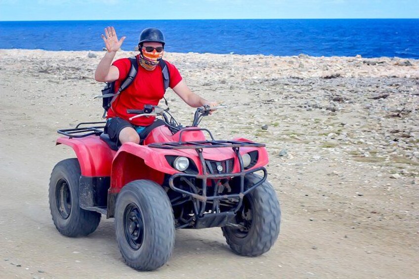 Eine Person fährt mit einem roten Quad auf einem unbefestigten Weg in der Nähe eines felsigen Strandes, im Hintergrund das Meer. 1