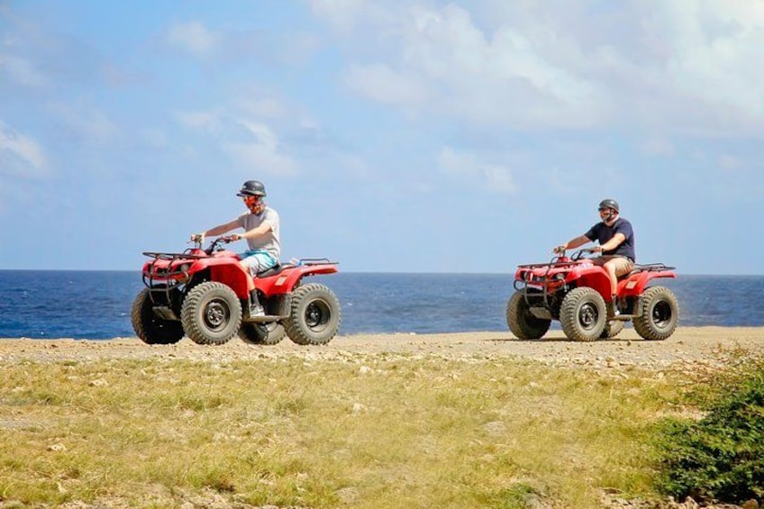 Two people riding red ATVs on a coastal road with the sea in the background. 2