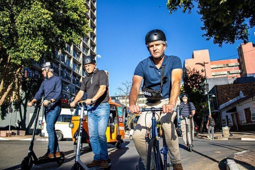 Three people riding scooters on a city street with buildings and a bus in the background. 2