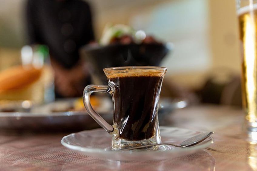 A clear glass cup of coffee on a saucer with a spoon, a blurred background of a person and a bowl of food. 5
