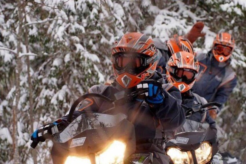 Two riders on a snowmobile, wearing helmets and gloves, in a snowy forest. 3