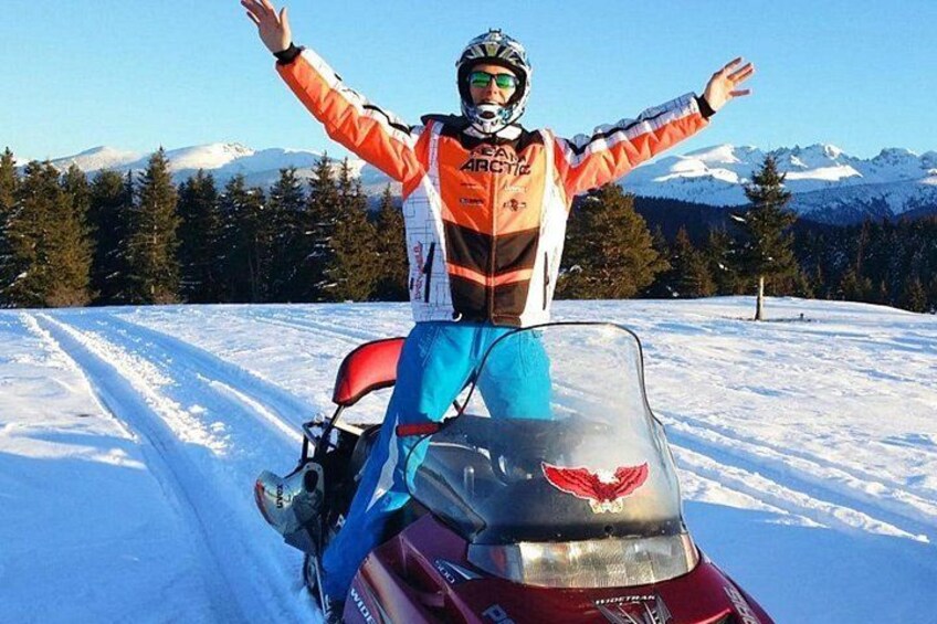 A person on a snowmobile with arms outstretched, wearing a helmet and goggles, on a snowy trail with mountains in the background. 2