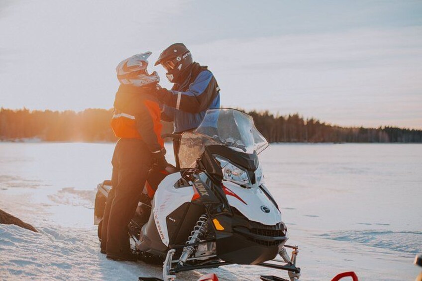 Two people on a snowmobile, wearing helmets and jackets, on a frozen lake with trees in the background. 2
