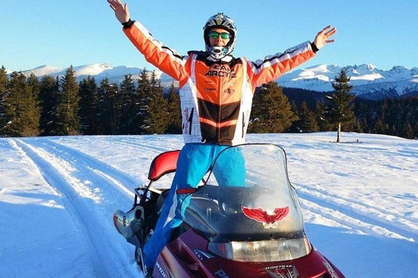 A person on a snowmobile with arms outstretched, wearing a helmet and goggles, on a snowy path with trees and mountains in the background. 4