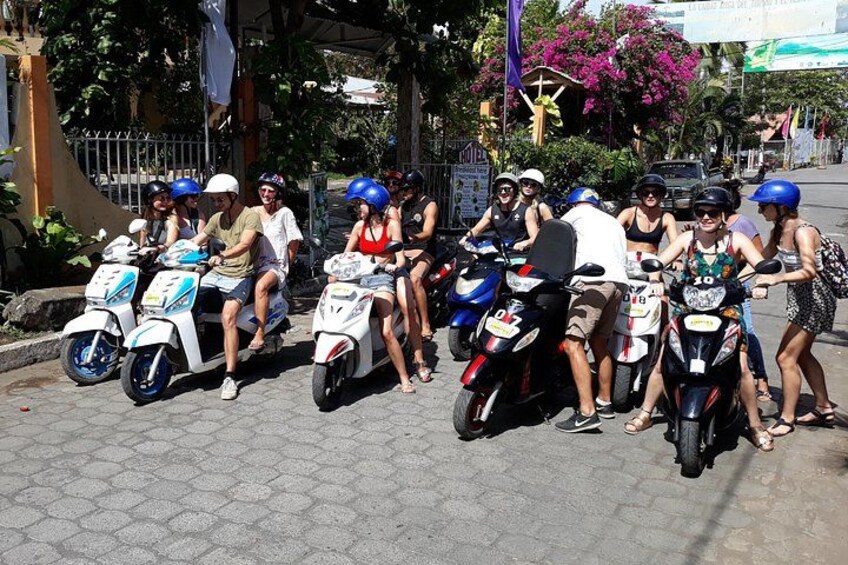 A group of people on scooters, some wearing helmets, on a paved street with trees and buildings in the background. 3