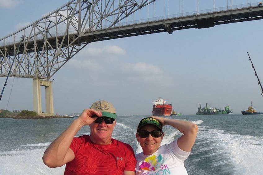 Two people on a boat with a bridge and cargo ships in the background. 4