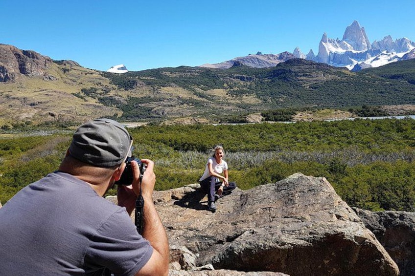 A person taking a photograph of another person sitting on a rock with a mountainous landscape in the background. 4