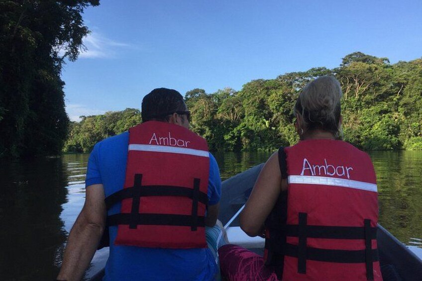 Two people in a canoe wearing red life jackets with the word 'Ambar' on them, paddling on a calm river surrounded by dense green foliage. 3
