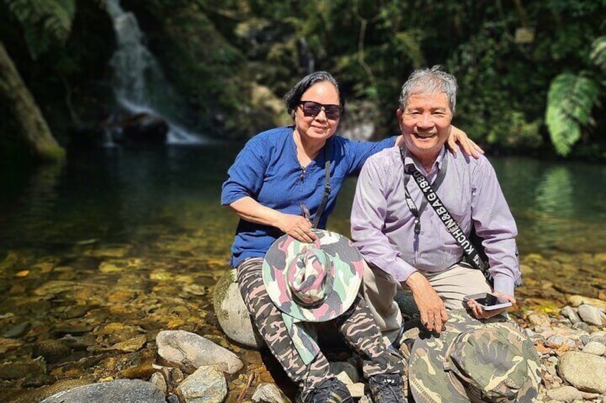 Zwei Personen sitzen auf Felsen an einem klaren Bach, im Hintergrund ein Wasserfall. 1