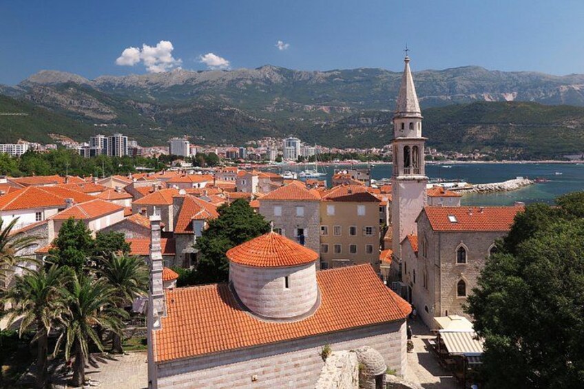 A coastal cityscape with red-roofed buildings, a prominent church tower, and a mountainous backdrop. 5