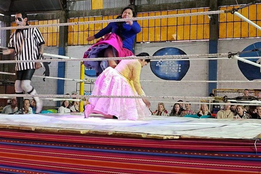 En brottningsmatch i en ring med en domare och två brottare, varav den ena har en blå överdel och ett rosa kjol, och den andra har en gul överdel och en rosa kjol. 3